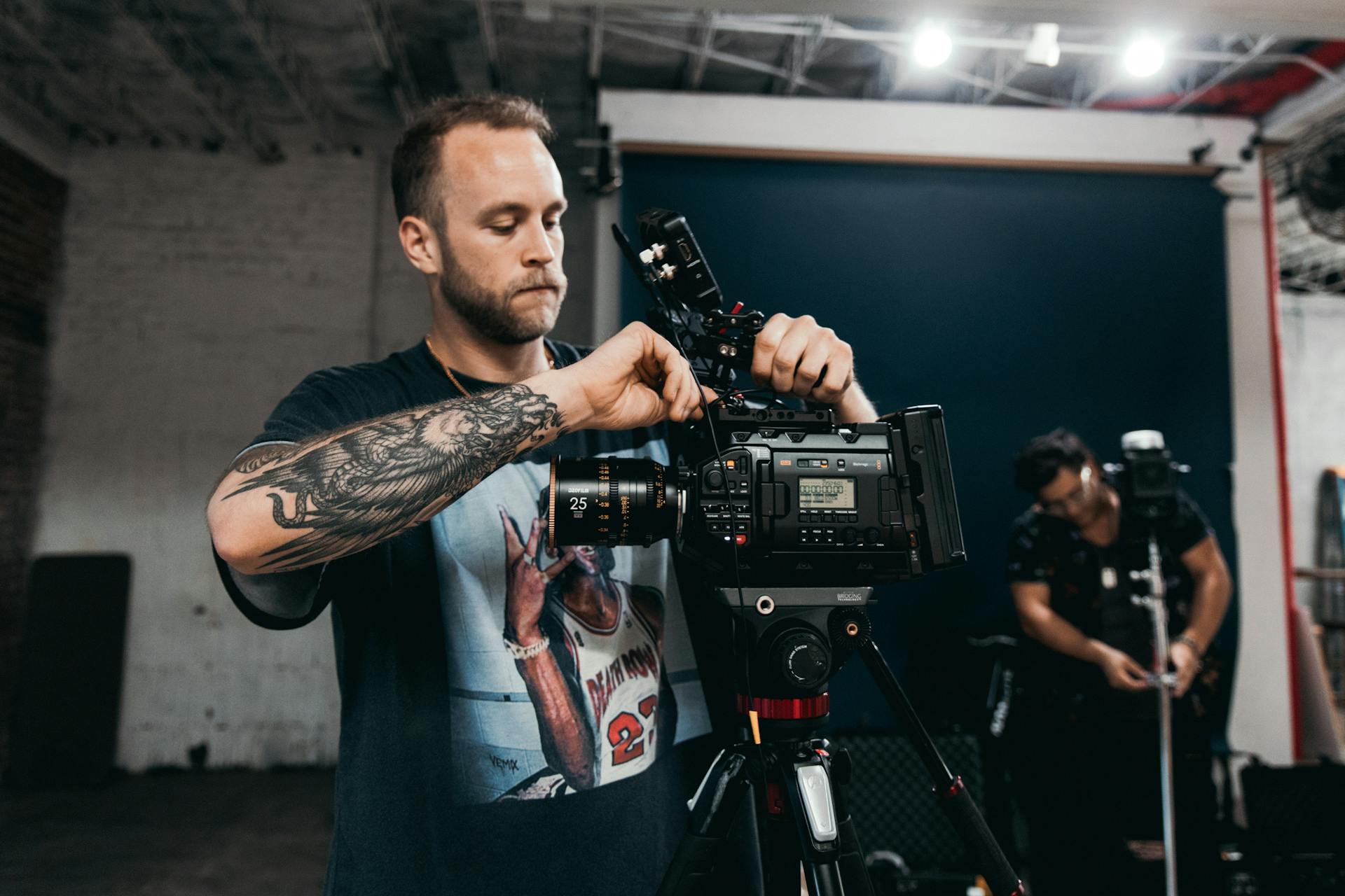 A bearded videographer adjusts a cinema camera on a tripod in a professional studio environment.