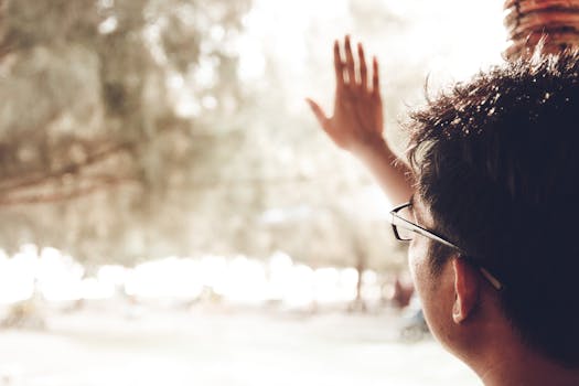 A man wearing glasses waves his hand outdoors, captured in bright daylight.