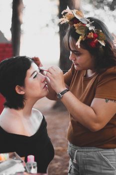 A makeup artist carefully applies eyeshadow to a woman's eyes in an outdoor setting.