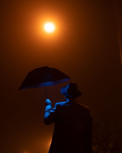 Free Silhouette of a man with an umbrella against a foggy, lit night sky. Stock Photo