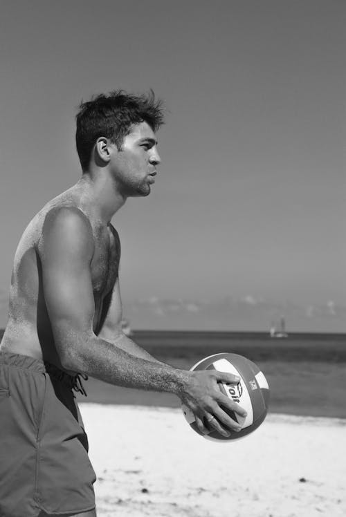 Grayscale Photo of a Shirtless Man Holding a Volleyball at the Beach