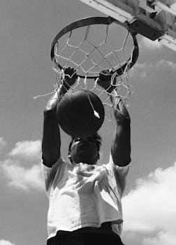Monochrome image capturing an intense basketball dunk against a clear sky.