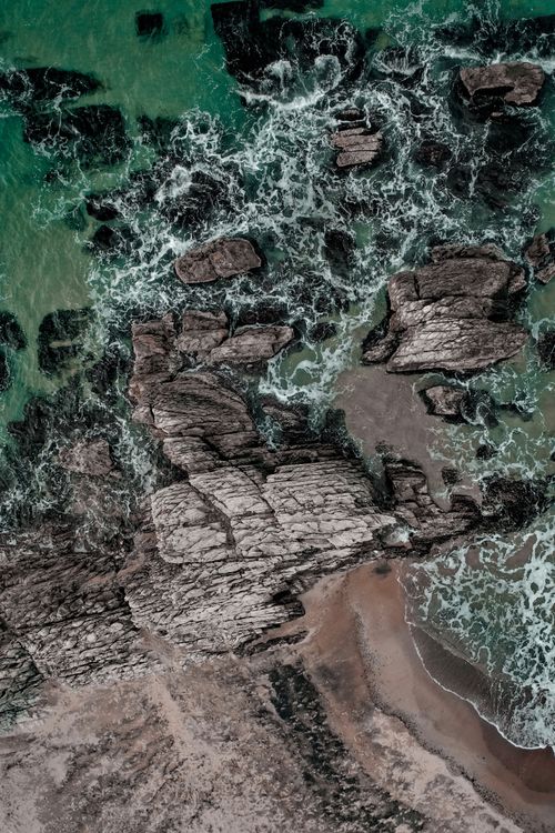 Free Stunning aerial view of rocky shoreline with waves crashing onto the rocks beneath clear skies. Stock Photo