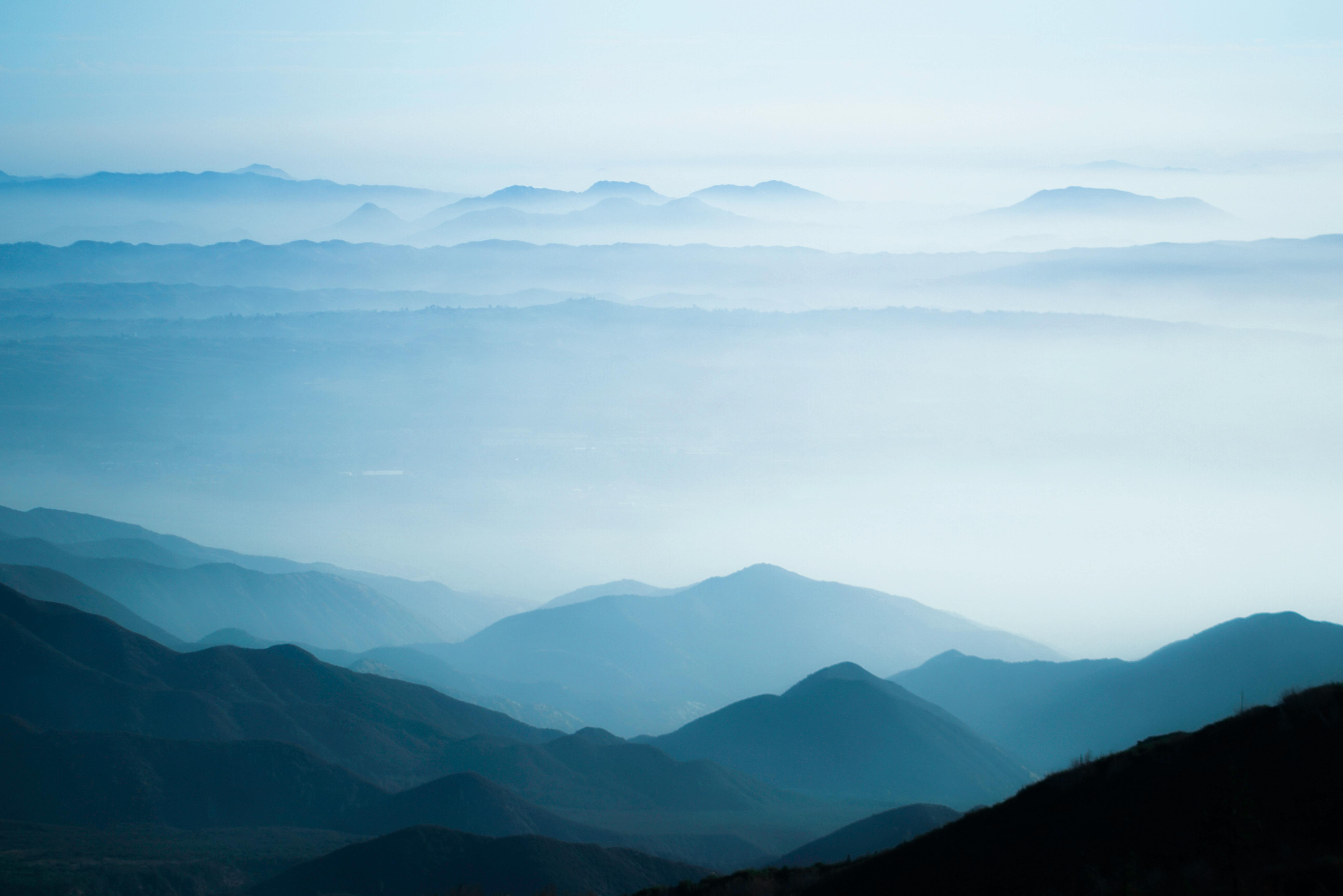 A tranquil scene of misty mountain ranges under a soft, blue sky at sunrise.