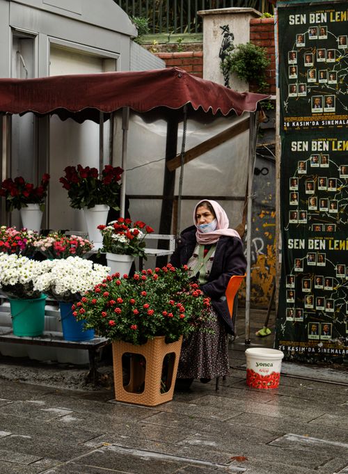 Free An elderly woman wearing a mask sits beside her flower stand on a wet urban street. Stock Photo