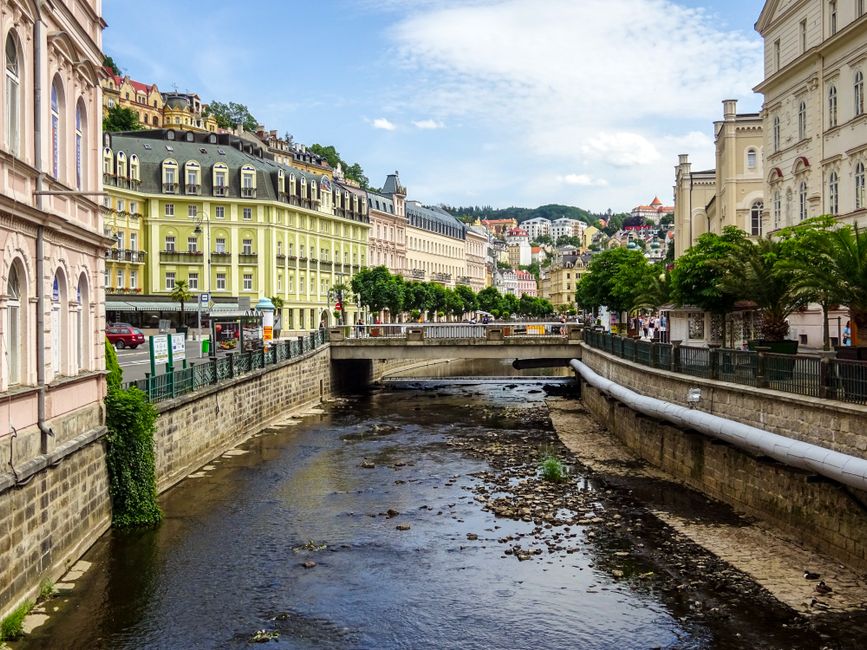 Scenic view of Karlovy Vary's canal with historic buildings and lush greenery under a blue sky.
