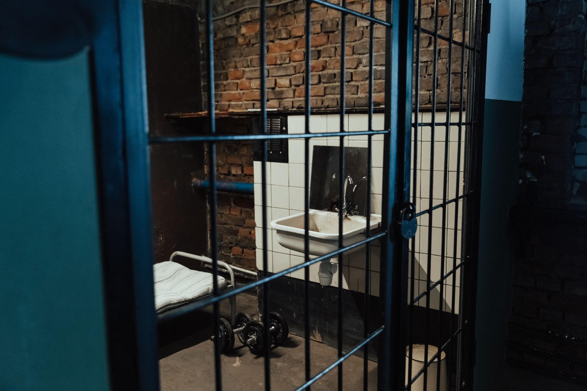 A dimly lit prison cell interior featuring a bed, sink, and exposed brick walls, evoking feelings of isolation.