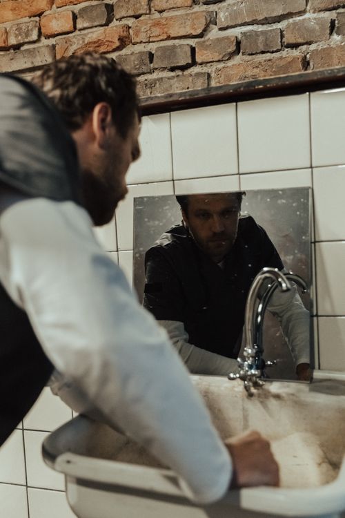 Free Man staring at mirror above an old bathroom sink in rustic setting, conveying introspective mood. Stock Photo