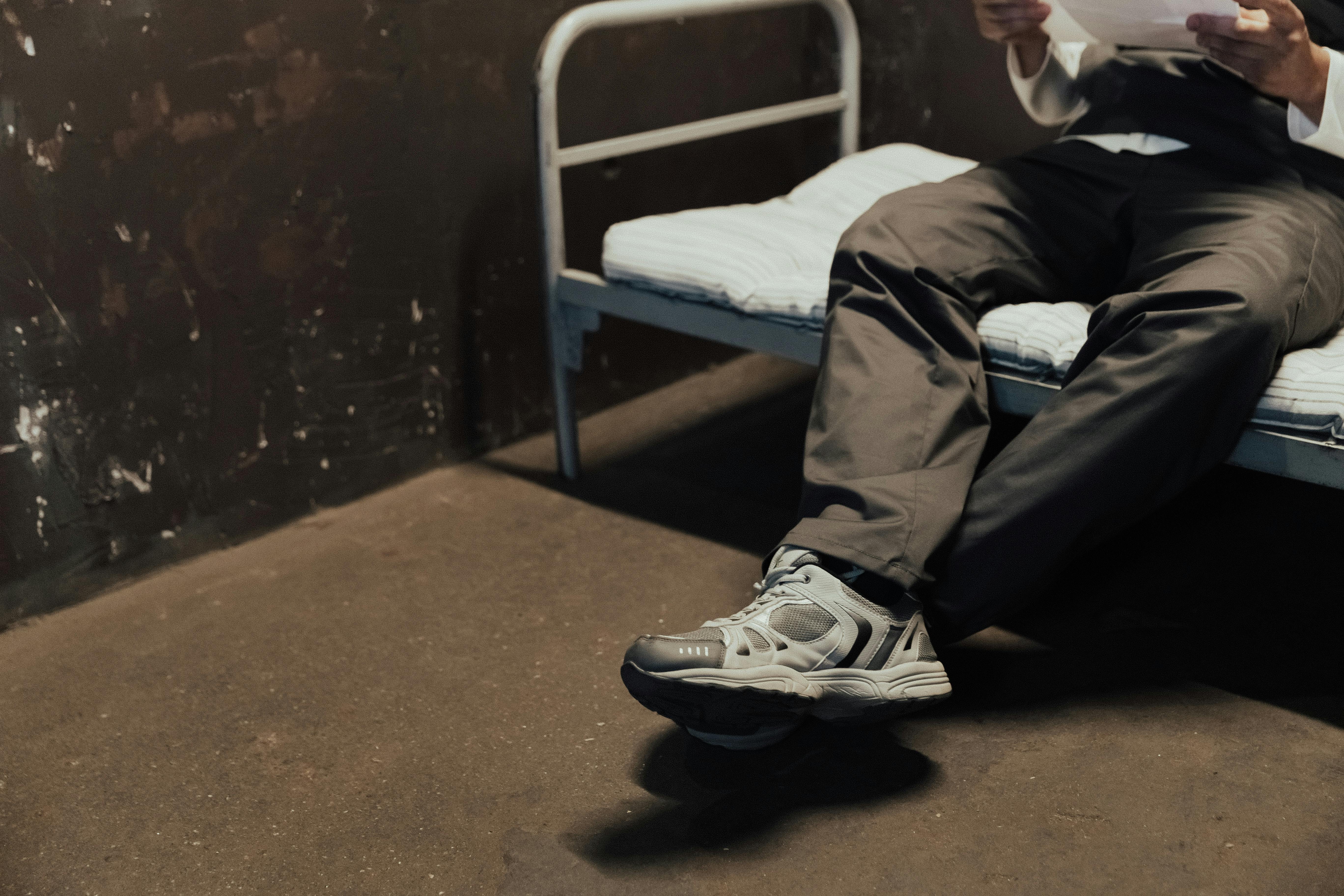 Man in casual attire sits on a simple bed in an industrial setting holding a paper.