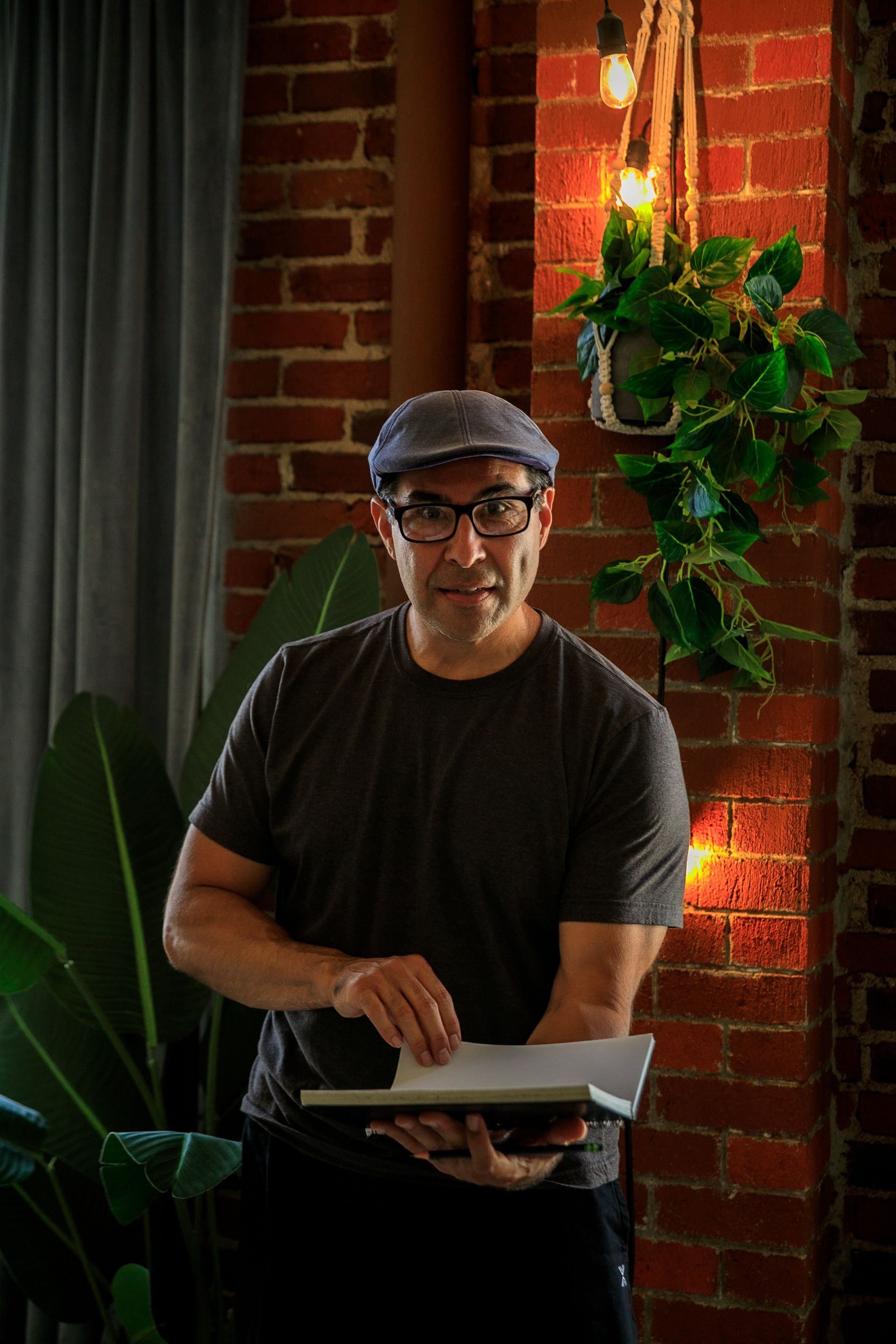 A man wearing a hat and glasses holding a sketchbook indoors with brick walls and greenery.