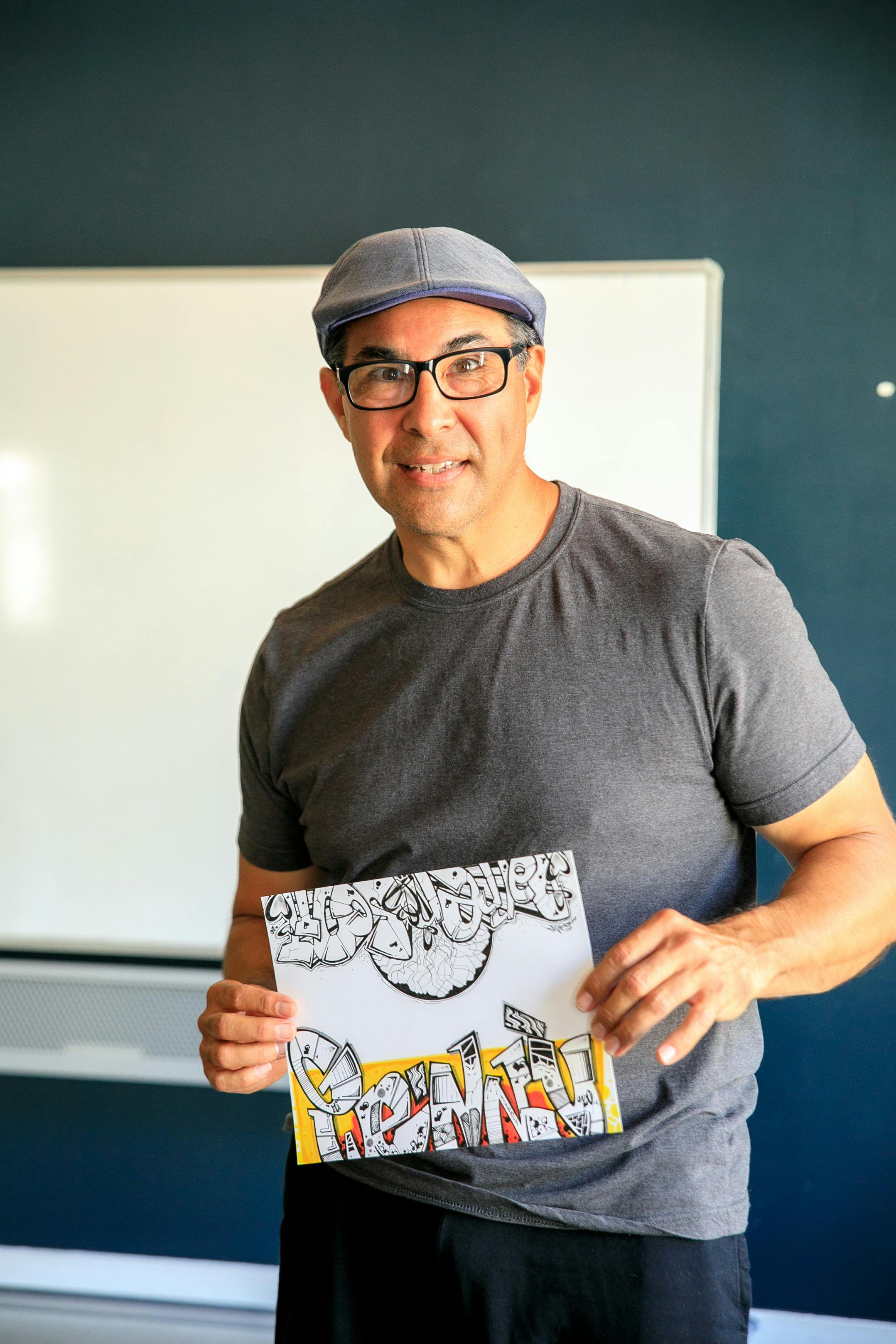 Man in a classroom presenting graffiti-inspired art, wearing a cap and glasses.