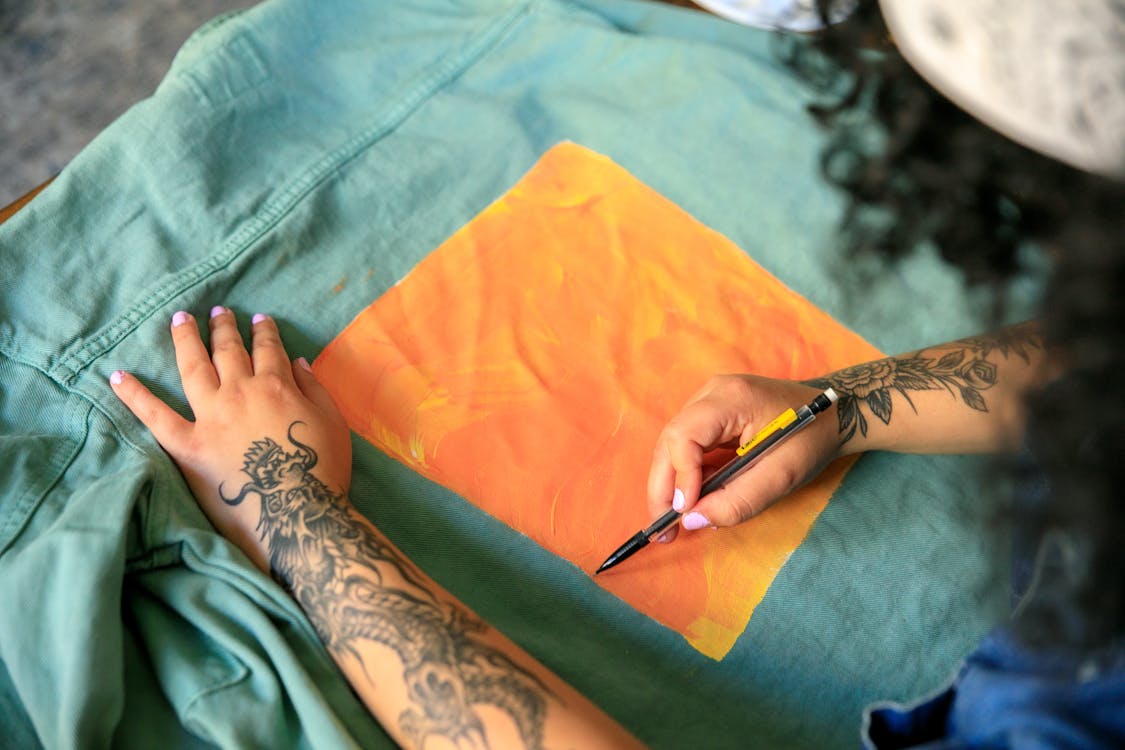 Free Tattooed artist adds bright orange artwork to a green jacket, showcasing creativity and design. Stock Photo