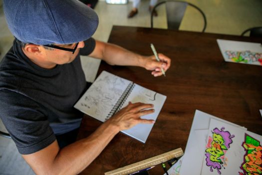 Top view of a man sketching graffiti art in a notebook at a wooden desk.