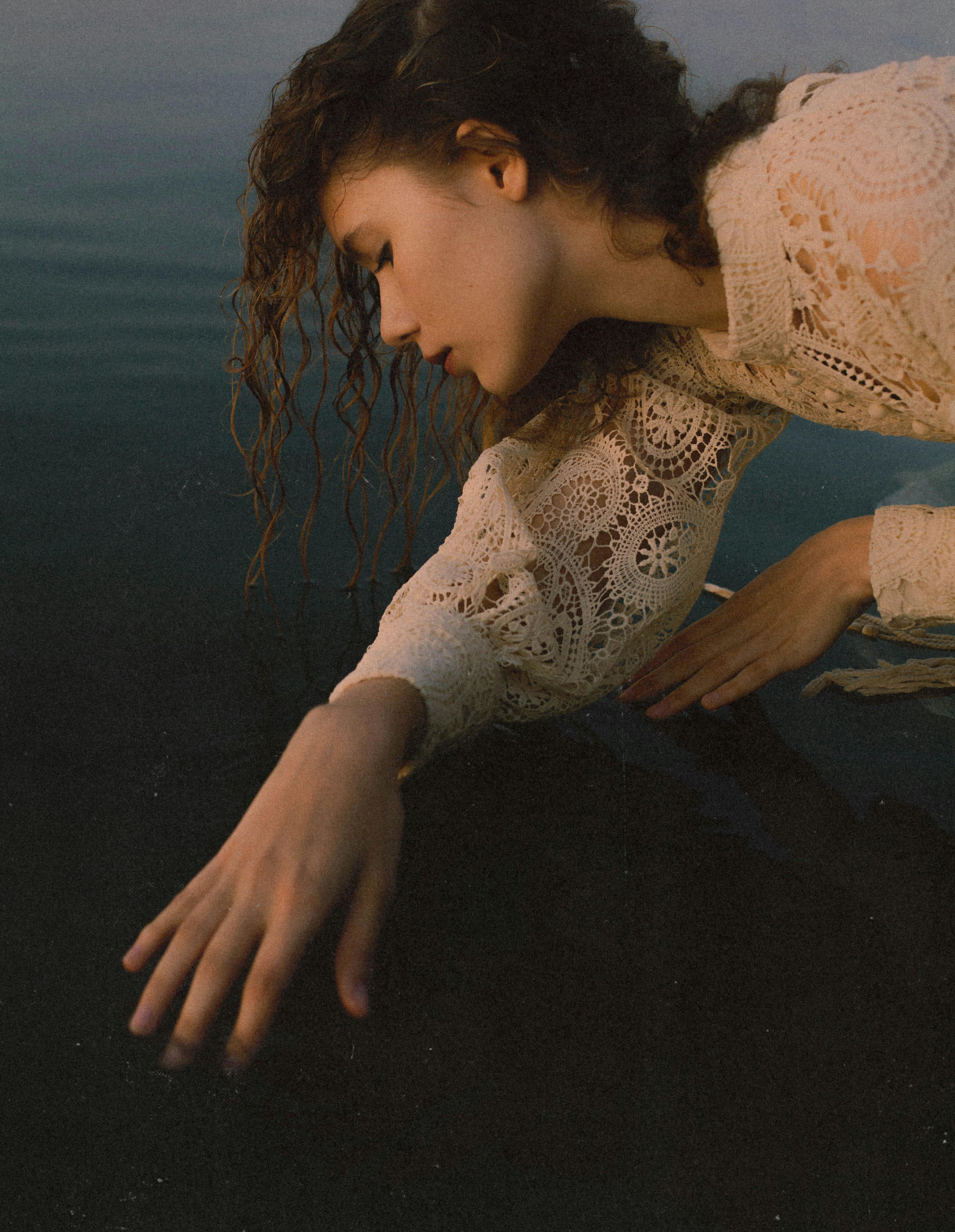 Woman in lace dress gently touching water surface in a serene moment.