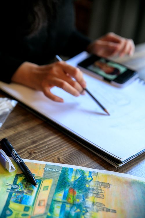 Free A woman sketching in a notebook with vibrant artwork nearby on a wooden table. Stock Photo