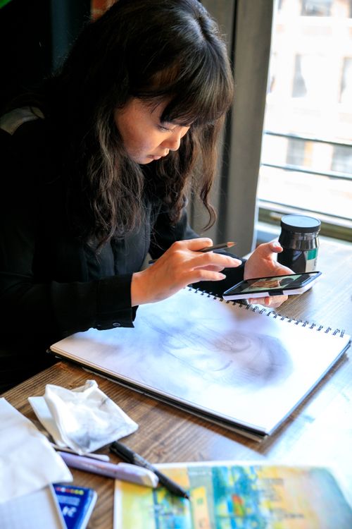 Free Artist using a smartphone for reference while sketching in a bright indoor setting. Stock Photo