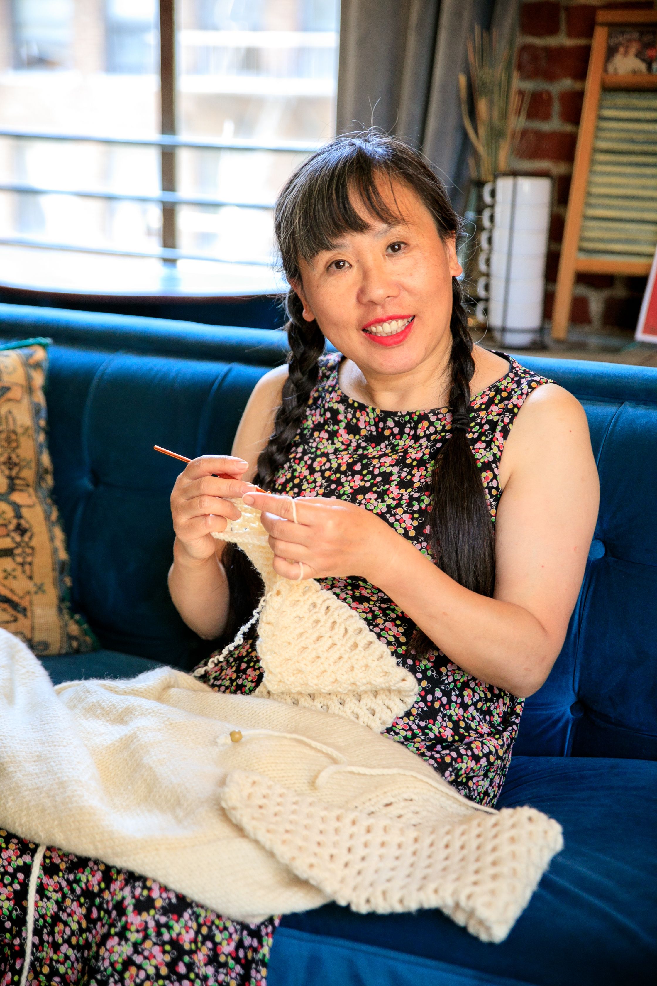 Smiling woman with black hair knitting on a blue couch indoors with bright daylight.