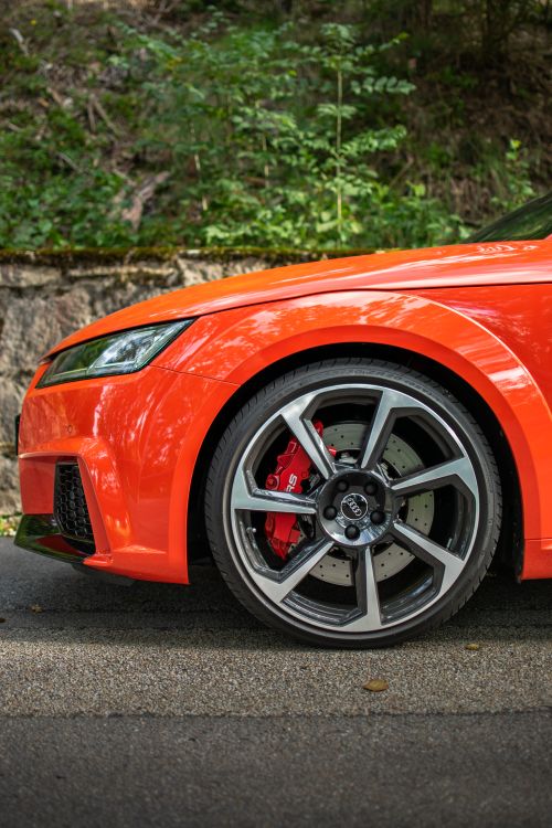 Free Detailed shot of a sleek orange car wheel on a sunlit road surrounded by greenery. Stock Photo