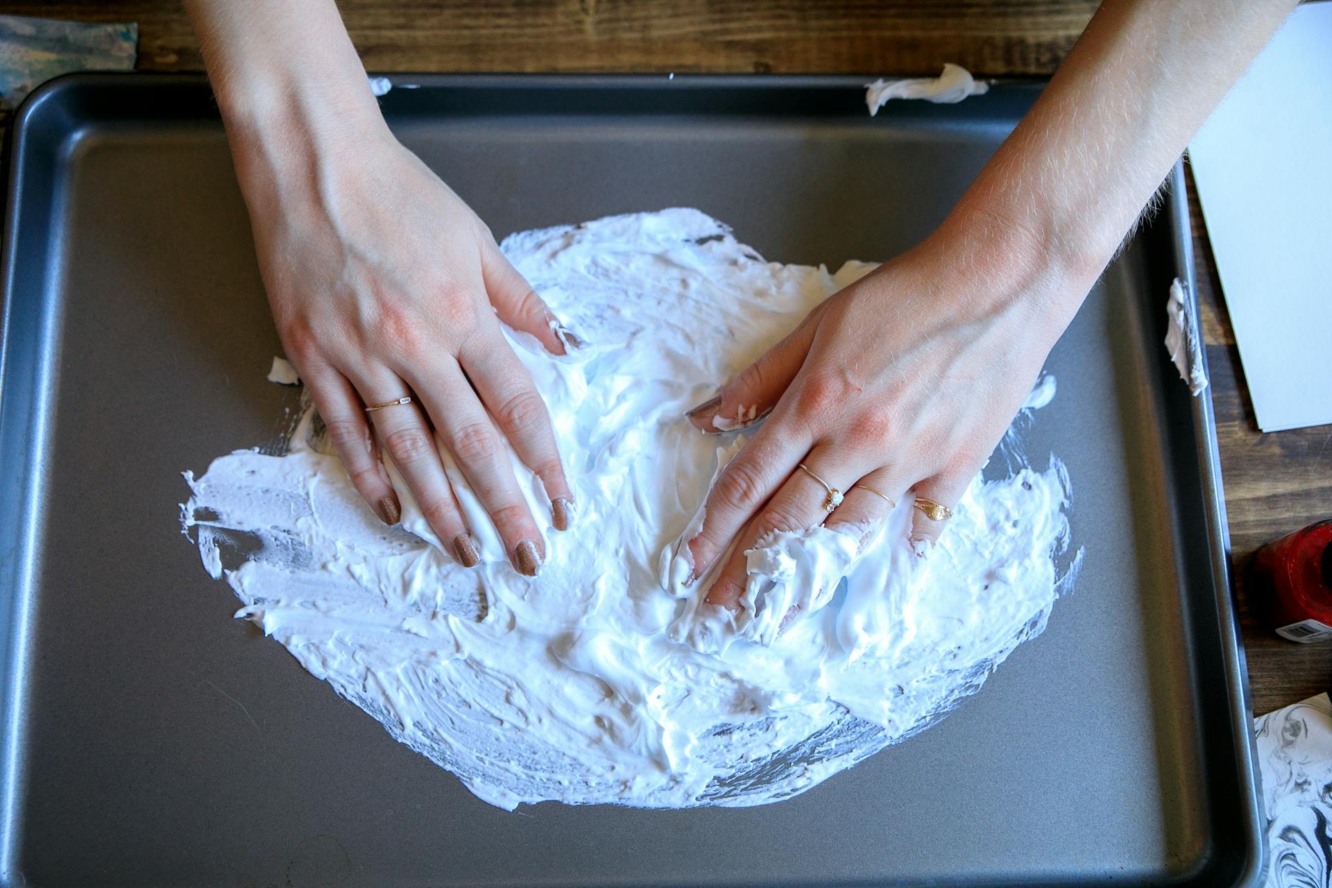 Hands spreading foam on a metal tray, creating texture and design, suitable for art or DIY projects.