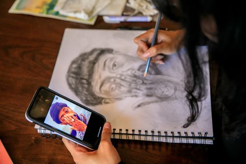 Free A person sketching a portrait using a smartphone reference on a spiral notebook. Stock Photo