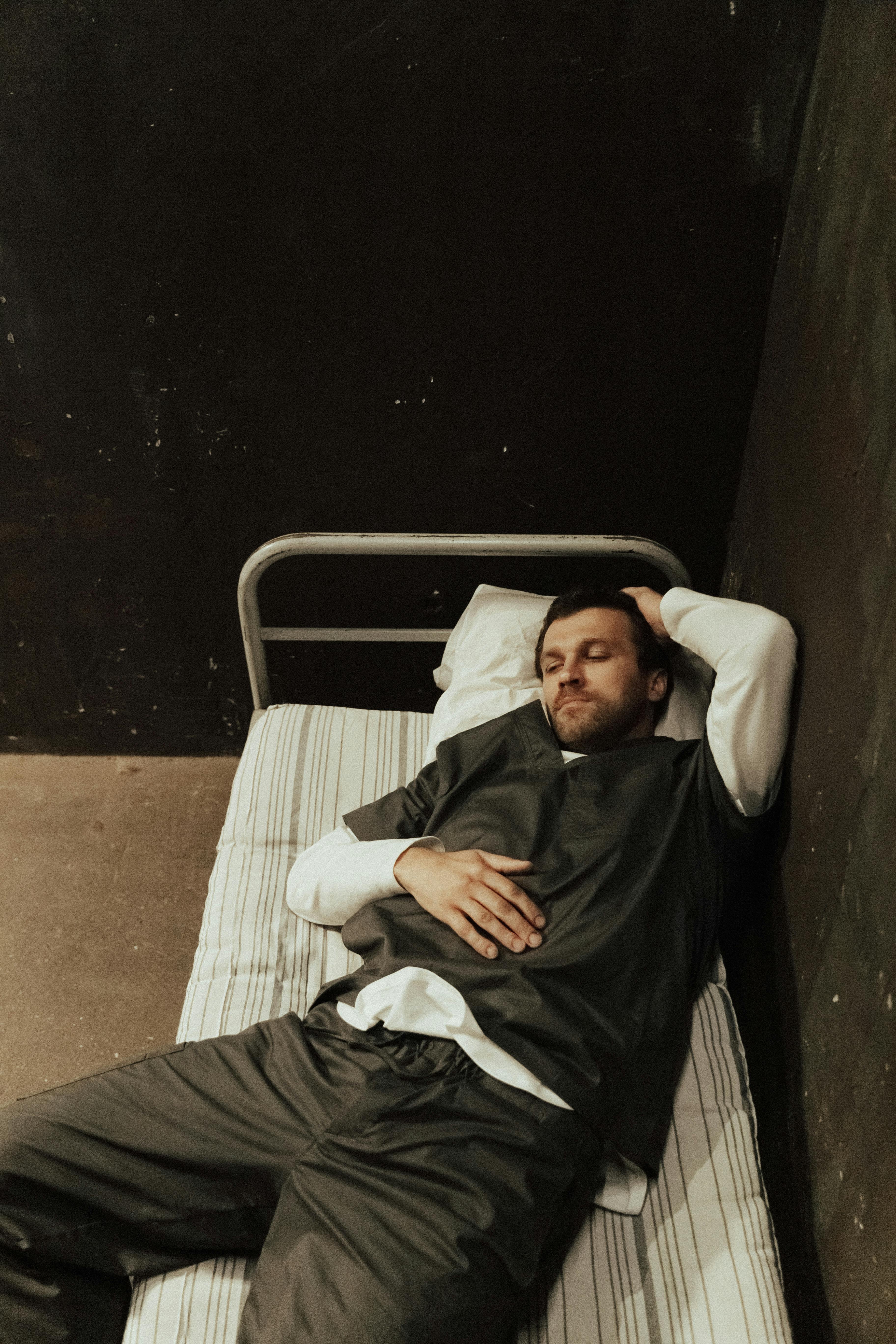 man in black shirt and white long sleeves lying on a bed near black walls