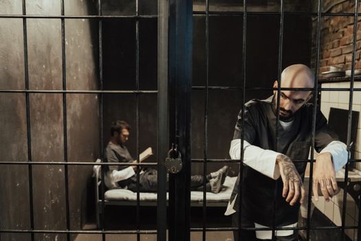Two men in a dimly lit prison cell, one reading on bed, the other standing somberly by bars.