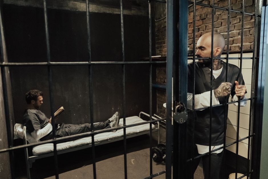 Two inmates in a prison cell, one reading and the other standing by the bars. Captures the essence of incarceration.