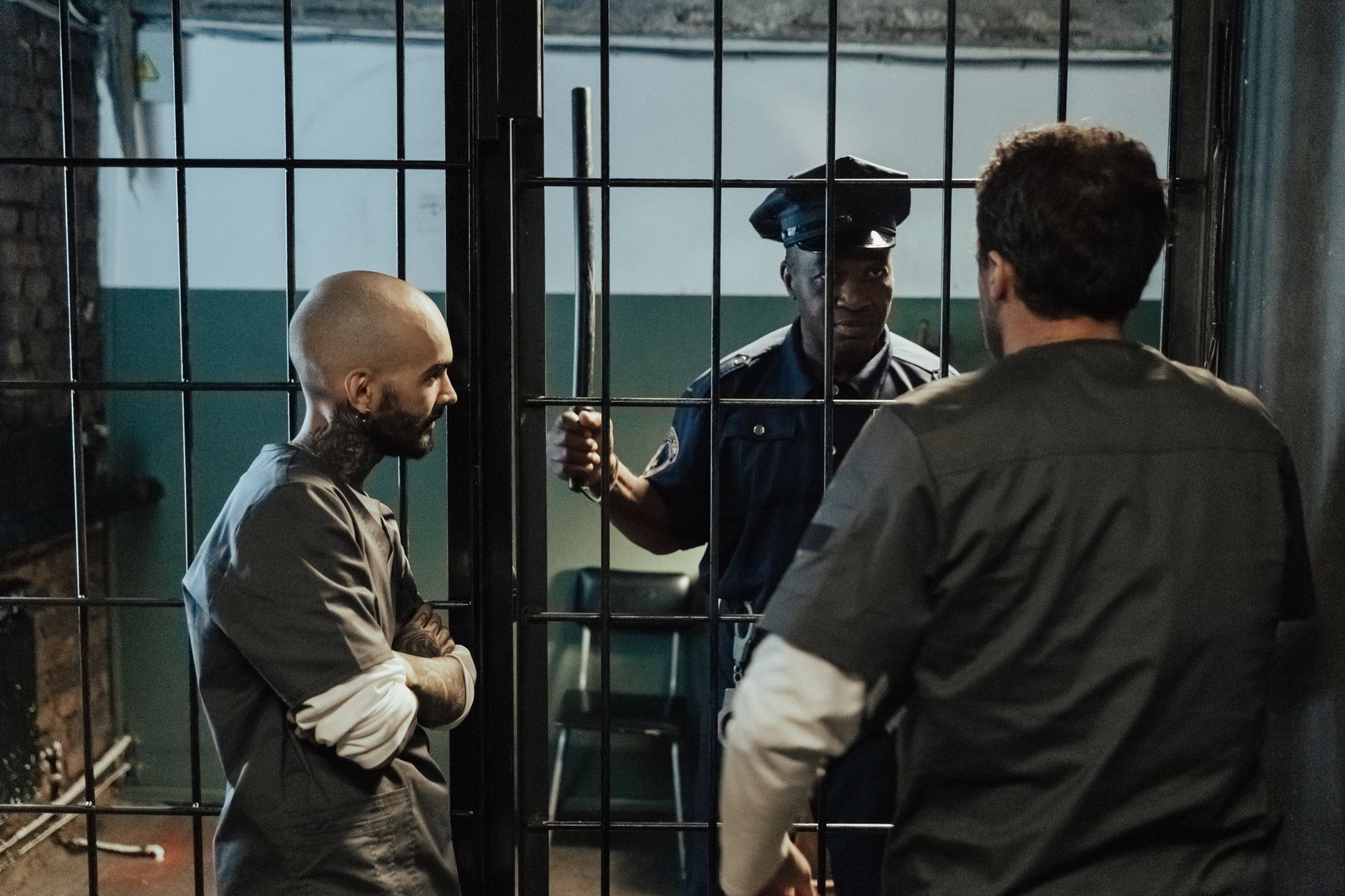 A prison guard interacts with two inmates inside a dimly lit jail cell.