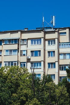 A beige apartment building surrounded by lush green trees under a clear blue sky.