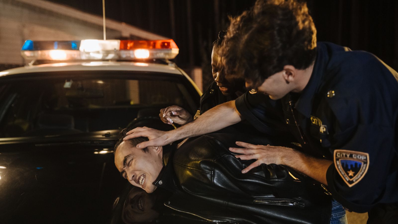 Dramatic nighttime arrest scene with police officers detaining a suspect on a patrol car.