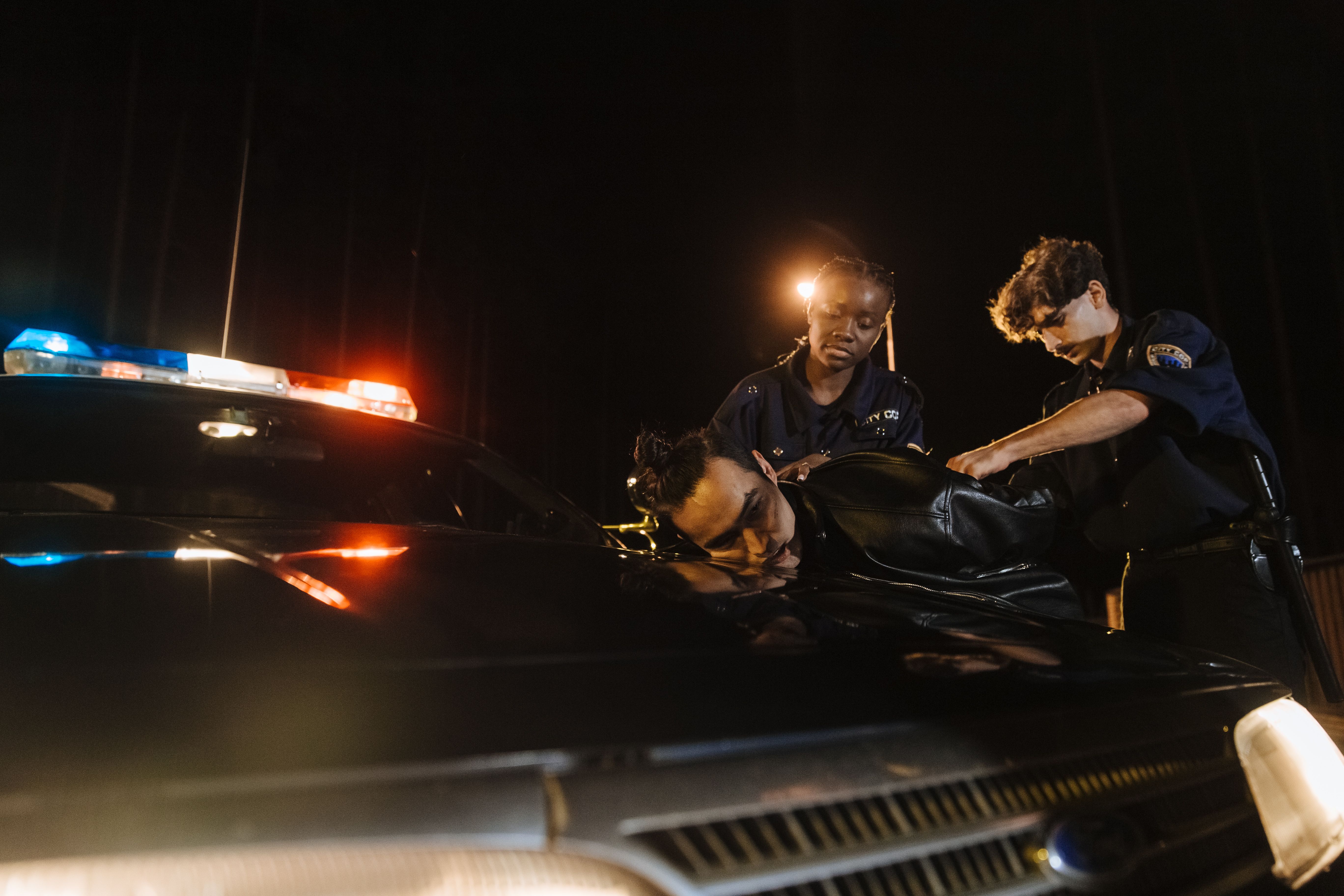 Two police officers restrain a man against a patrol car at night as its emergency lights flash