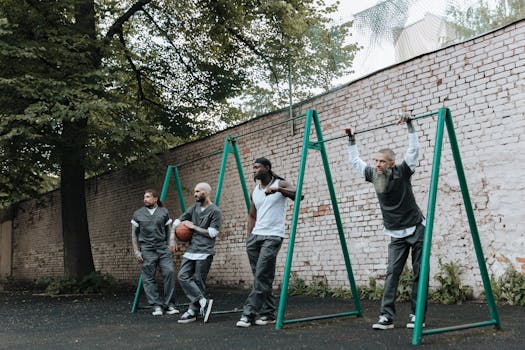 Men in casual attire playing basketball outdoors by a brick wall and exercise equipment.
