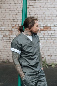 Tattooed man in scrubs leaning against a green pole outside near a brick wall.