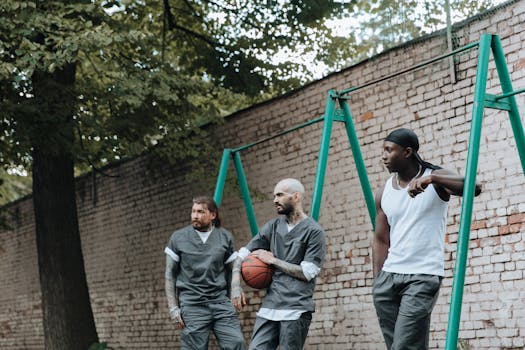 Three men in a prison yard, one holding a basketball, alongside a brick wall.