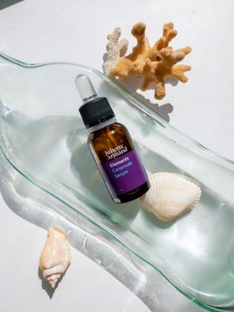 Serum bottle on glass with coral and seashells, top view.