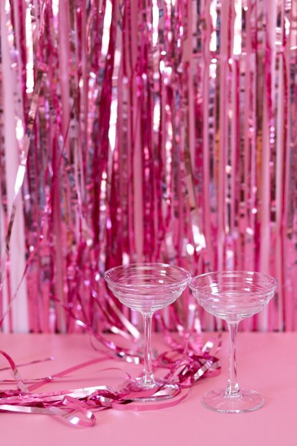 Two champagne glasses sit on a pink surface with a festive pink foil curtain backdrop.