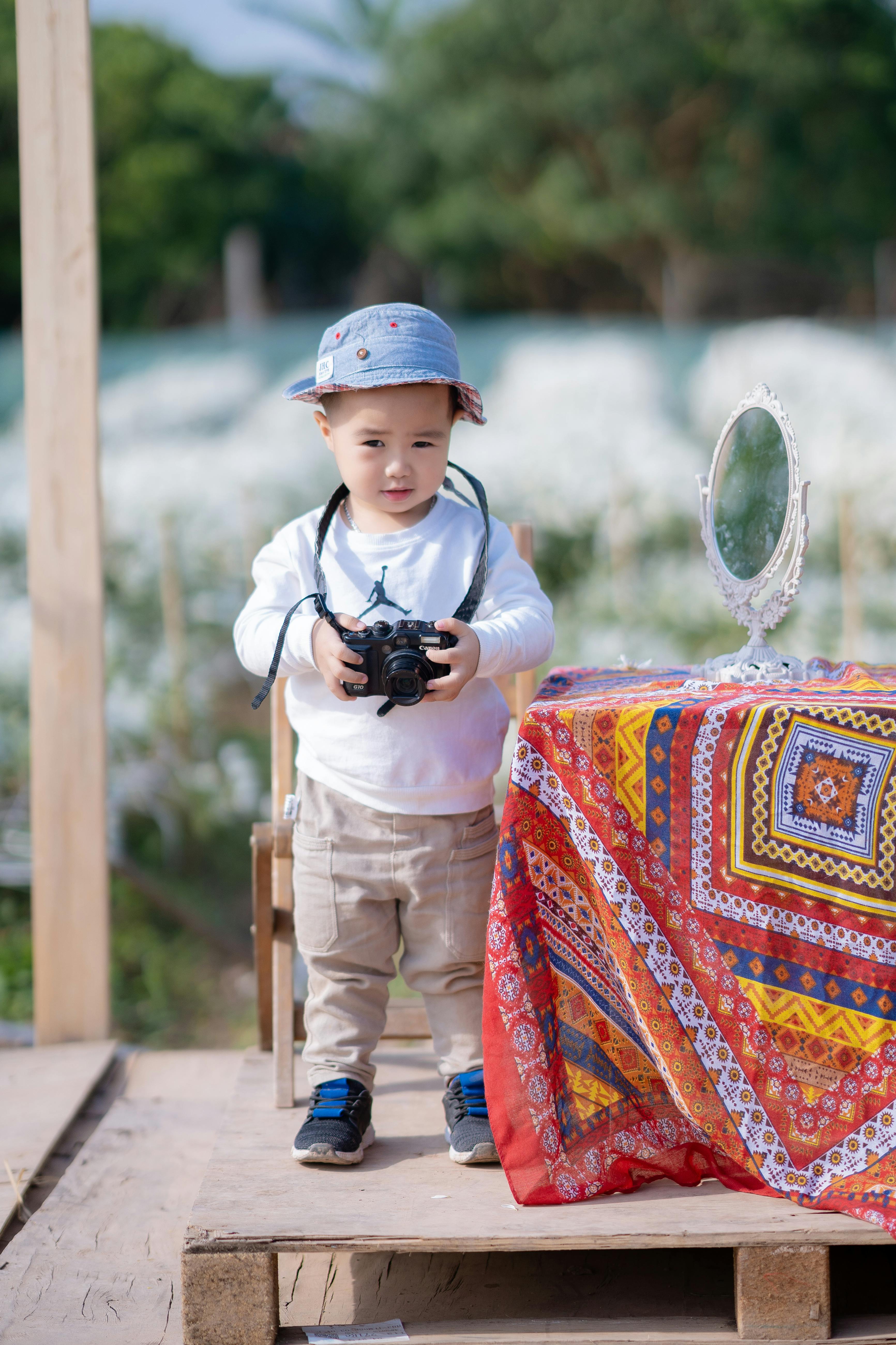 A young boy holding a camera, standing by a colorful tablecloth outdoors.