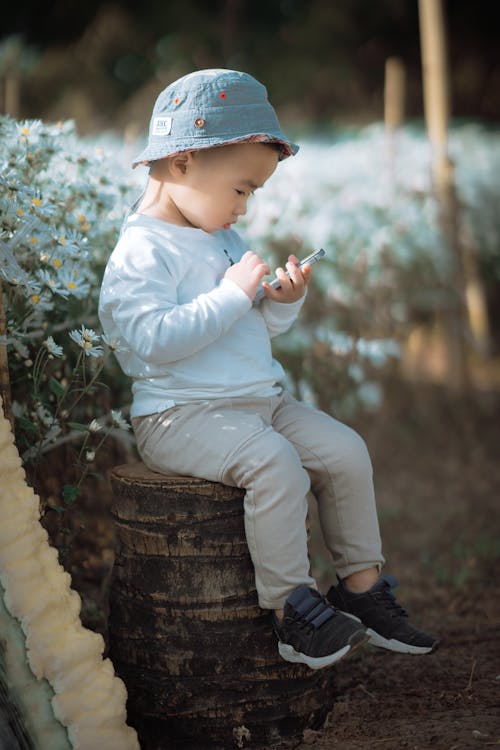 Free A Young Boy in White Sweater Sitting on a Tree Log while Holding a Mobile Phone Stock Photo