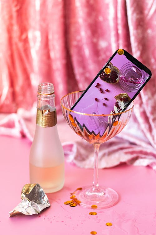 Smartphone Lying inside a Cocktail Glass and a Small Bottle of Champagne Standing against Pink Background 