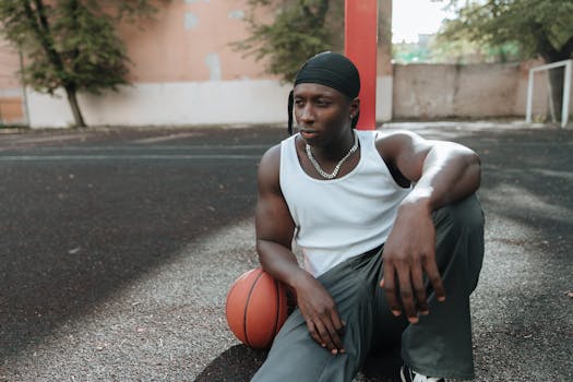 Young man sitting on outdoor basketball court, holding a basketball. Relaxed and thoughtful pose.