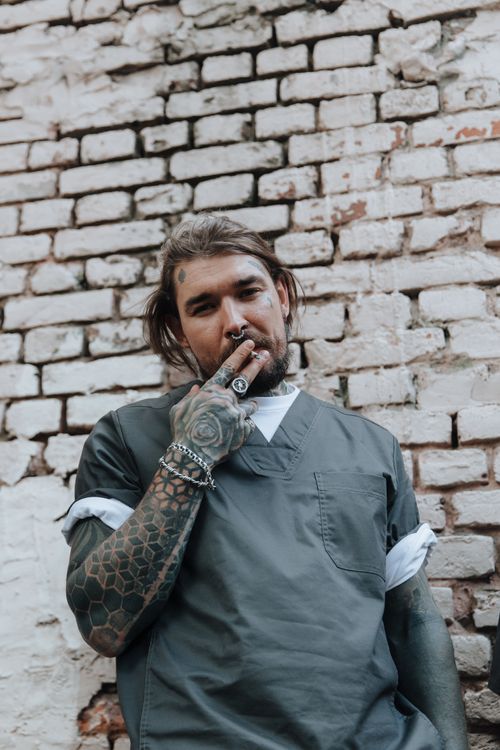 Free A tattooed man with a ponytail and piercings smokes outdoors against a brick wall. Stock Photo