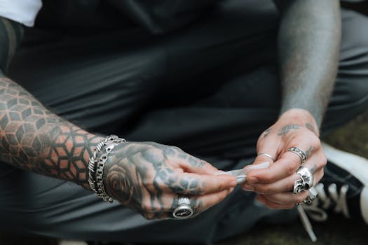 Close-up of tattooed hands rolling a cigarette, showcasing rings and bracelets on a relaxed outdoor setting.