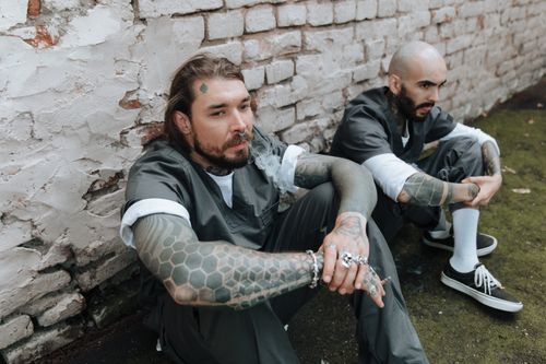 Free Two tattooed men in prison uniforms sit against a brick wall, exuding a gritty and rebellious vibe. Stock Photo