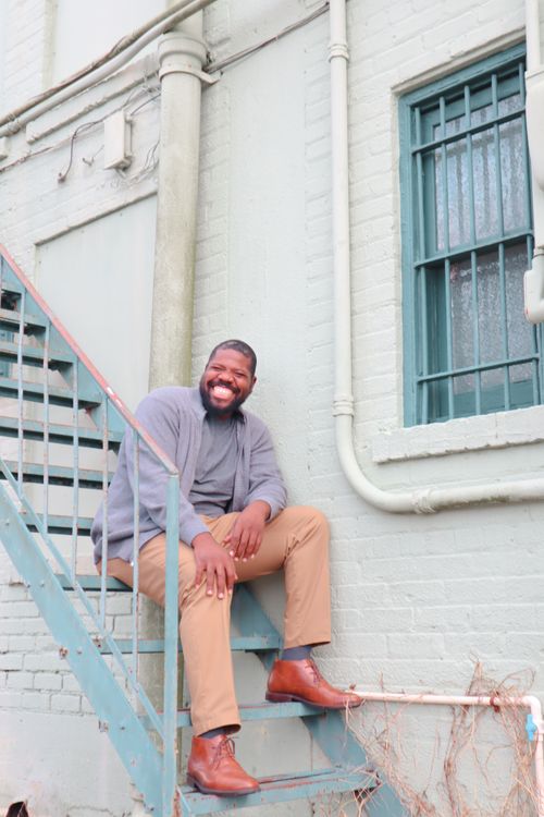 Free African American man with beard sitting and smiling on exterior metal stairs. Stock Photo