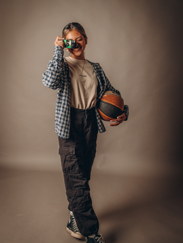 Woman in plaid shirt holds camera and basketball, smiling in studio setting.