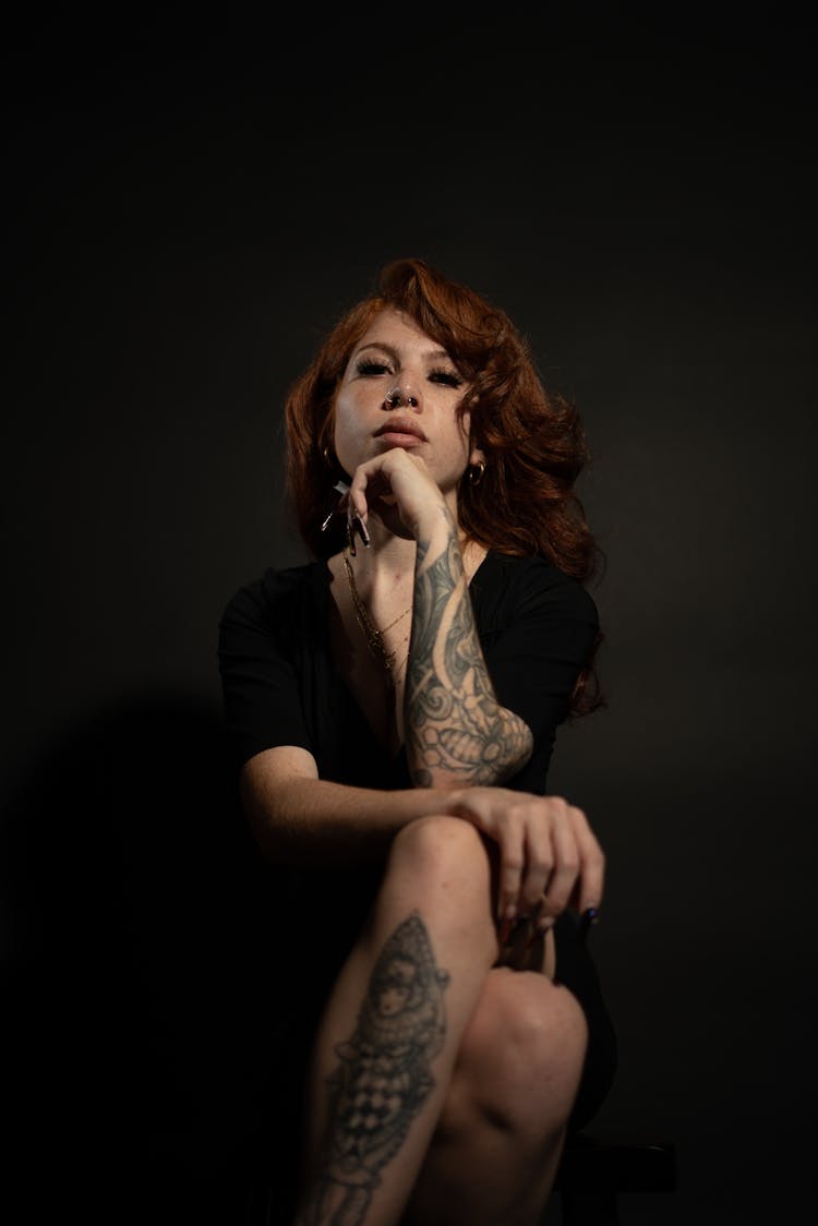 A Tattooed Woman In Black Shirt Sitting With Her Legs Crossed And Her Hand On Her Chin