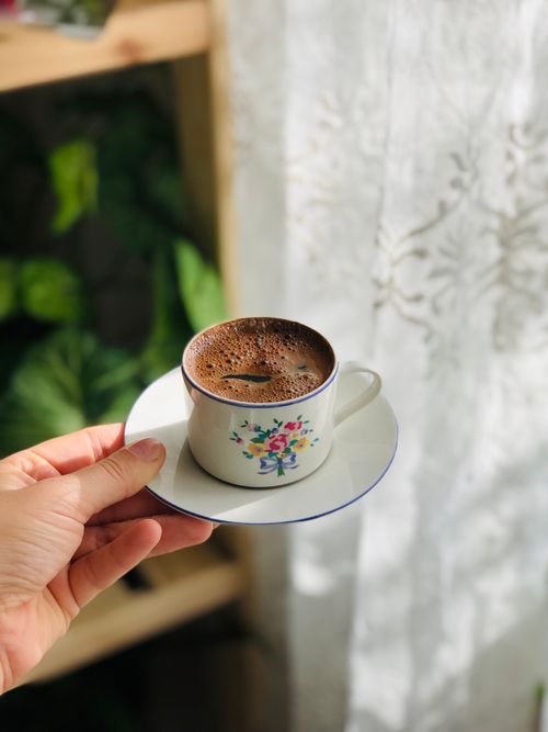 Free A hand holds a floral cup of Turkish coffee by a sunlit window, indoors. Stock Photo