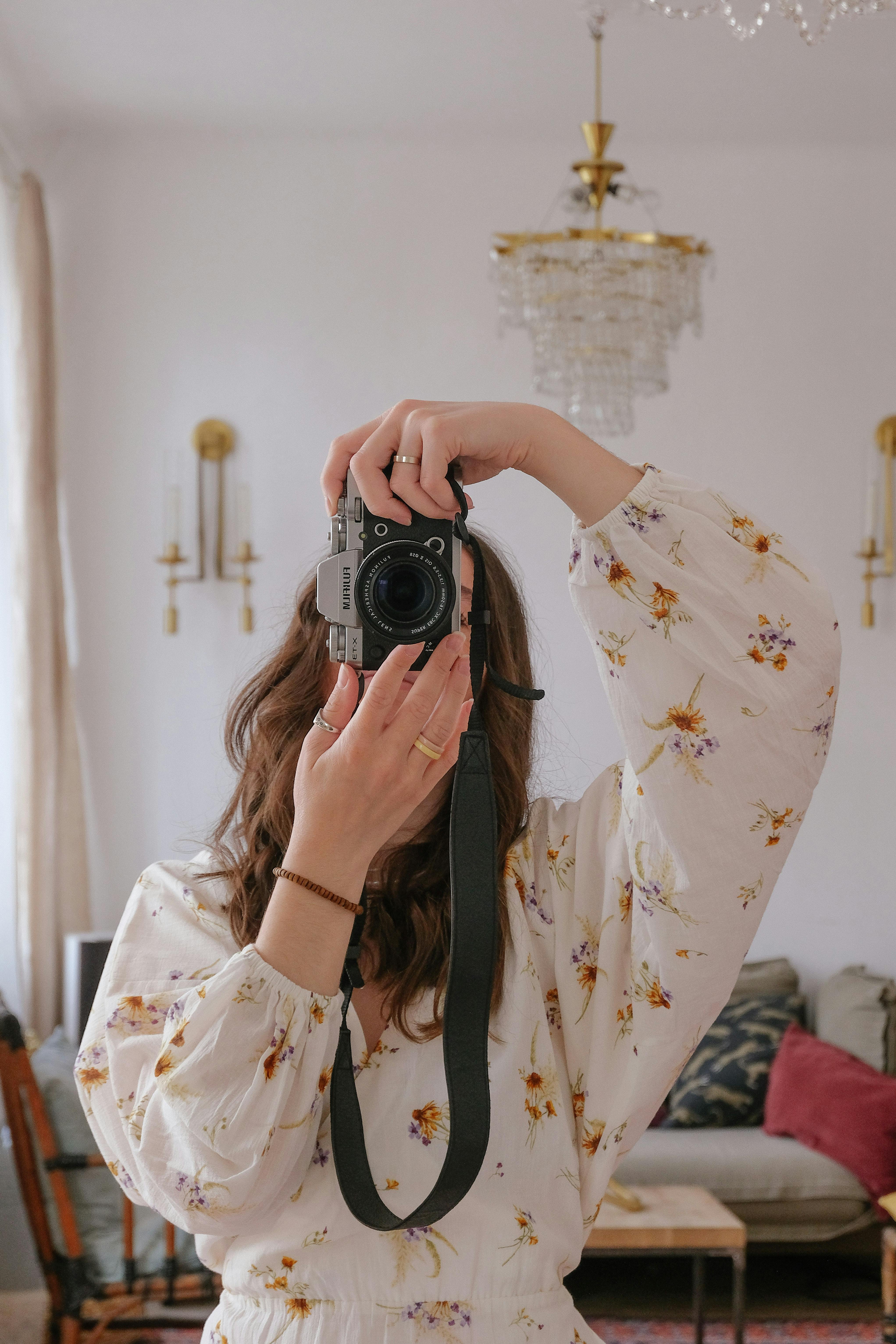 Free A woman in a floral dress photographs herself indoors with a DSLR camera. Stock Photo