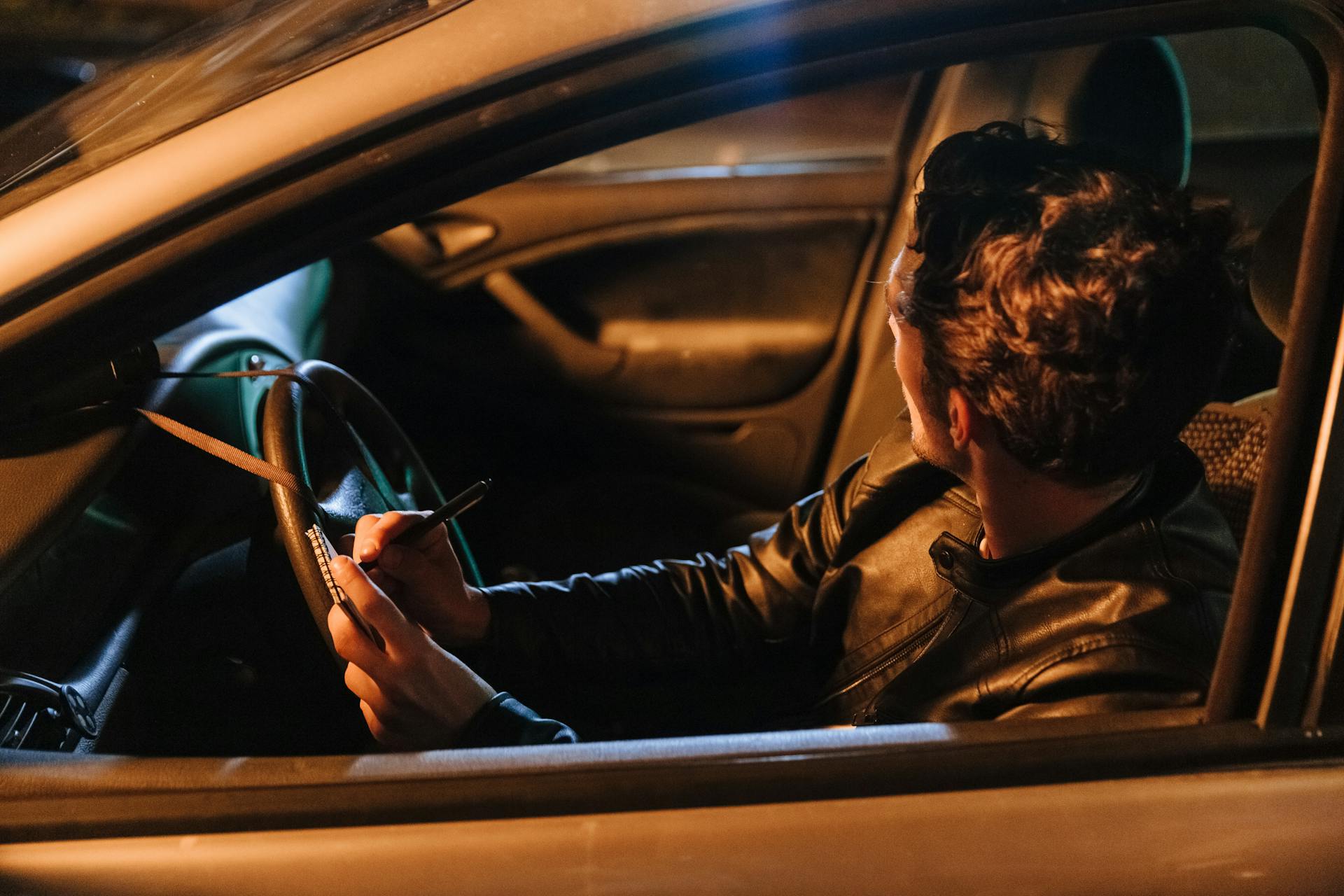 Man in Leather Jacket Sitting in a Car Taking Notes on a Stakeout