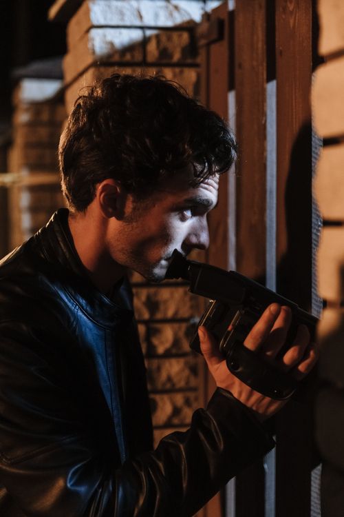 Free A man in a leather jacket holds a camera, investigating at night outdoors against a brick wall. Stock Photo